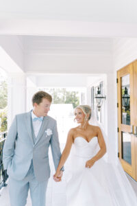 “Bride and groom walking hand-in-hand on a white porch, smiling at each other during their coastal summer wedding.”