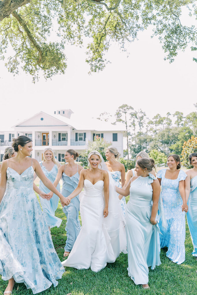 “A smiling bride walks hand-in-hand with her bridesmaids in light blue dresses on a sunny lawn in front of a white Southern-style mansion.”