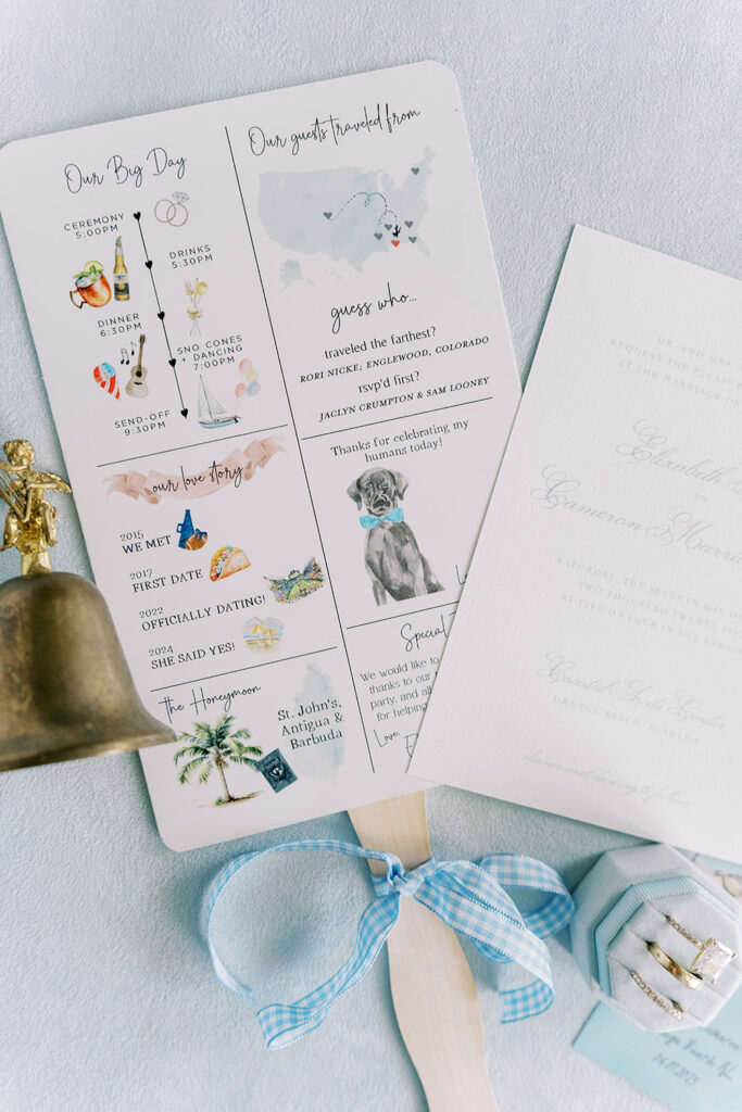 Wedding program fan decorated with illustrations of the couple’s love story, event timeline, and guest travel map, tied with a blue gingham ribbon, pictured next to wedding invitations, rings, and a bell.