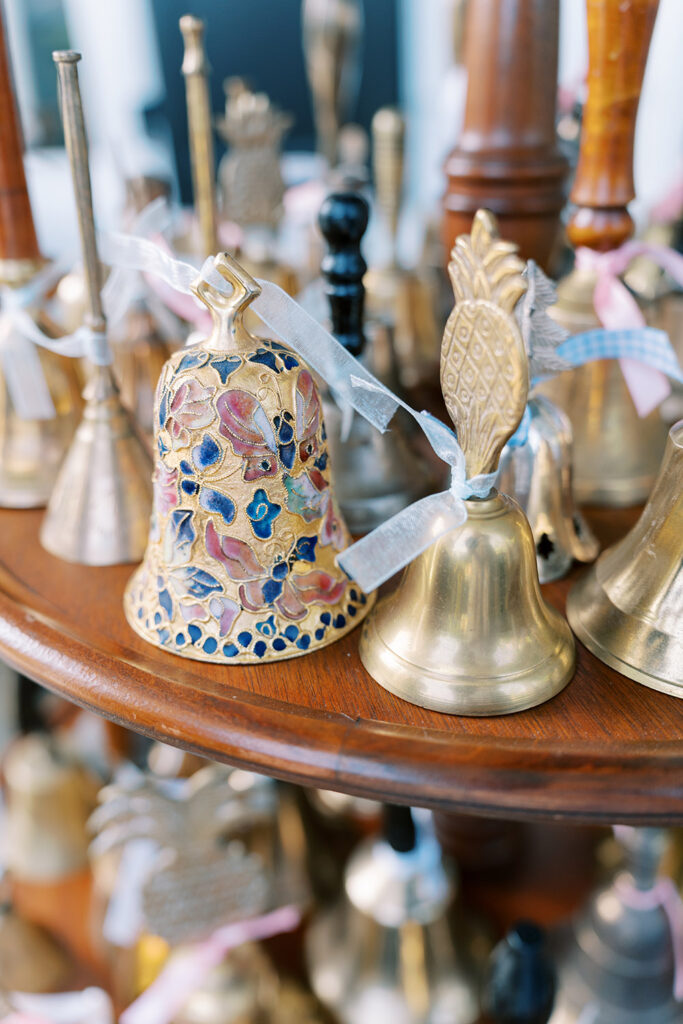 Close-up of a collection of small decorative bells on a wooden display, including a colorful enamel bell and a brass bell with a pineapple handle, each tied with a ribbon.