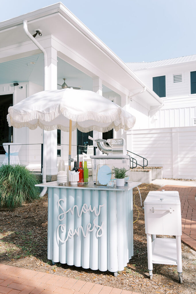 Outdoor snow cone cart at a wedding, featuring a light blue stand with “Snow Cones” lettering, colorful syrups, cups, and a white fringe umbrella in front of a white building.