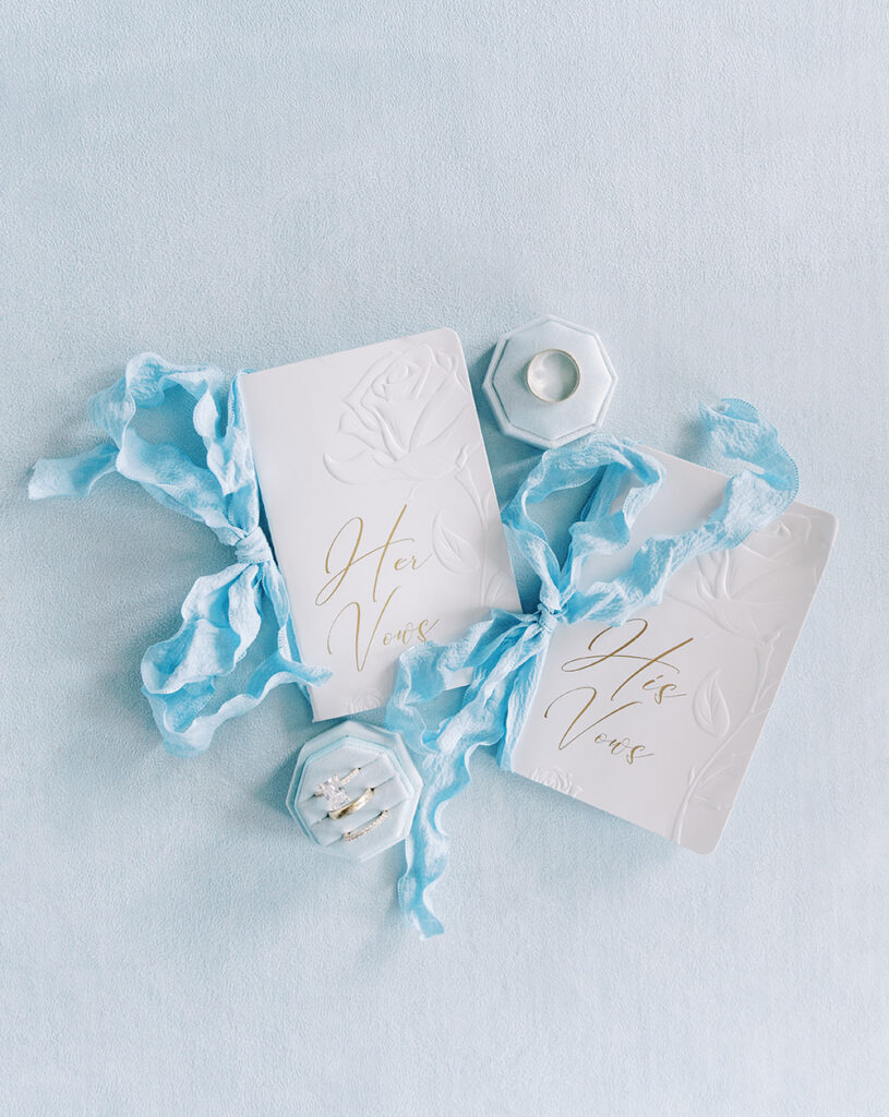“Elegant flatlay of white his and hers wedding vow booklets with blue ribbon, diamond rings, and wedding details on a soft blue background.”