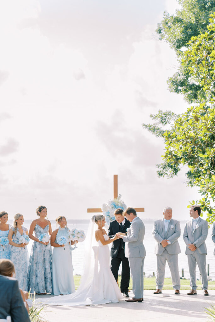 Bride and groom exchanging rings during an outdoor waterfront wedding ceremony, standing beneath a wooden cross decorated with blue and white flowers, surrounded by bridesmaids in light blue dresses and groomsmen in gray suits.