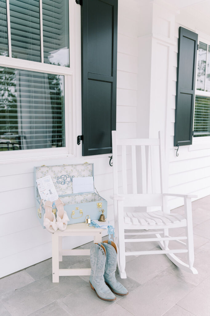 “Bridal details displayed on a white porch with a rocking chair, including blue cowboy boots, bow heels, a light blue suitcase with invitations, and a garter.”