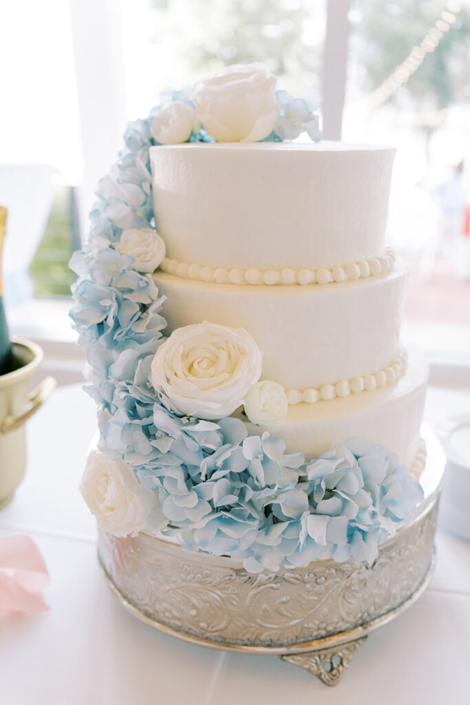 Close-up of a three-tier white wedding cake decorated with white roses and light blue hydrangeas cascading down the side, displayed on an ornate silver cake stand.