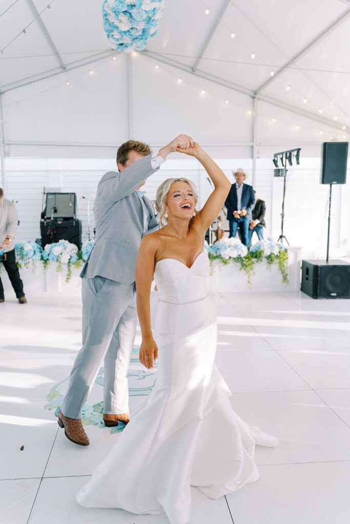 Bride and groom dancing during their wedding reception, smiling as the groom twirls the bride under a white reception tent decorated with string lights and blue floral arrangements.