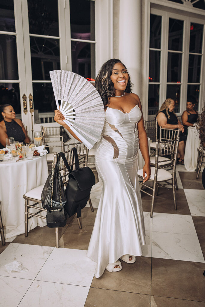 The bride smiles and holds a silver hand fan while walking through the reception in a fitted white gown.