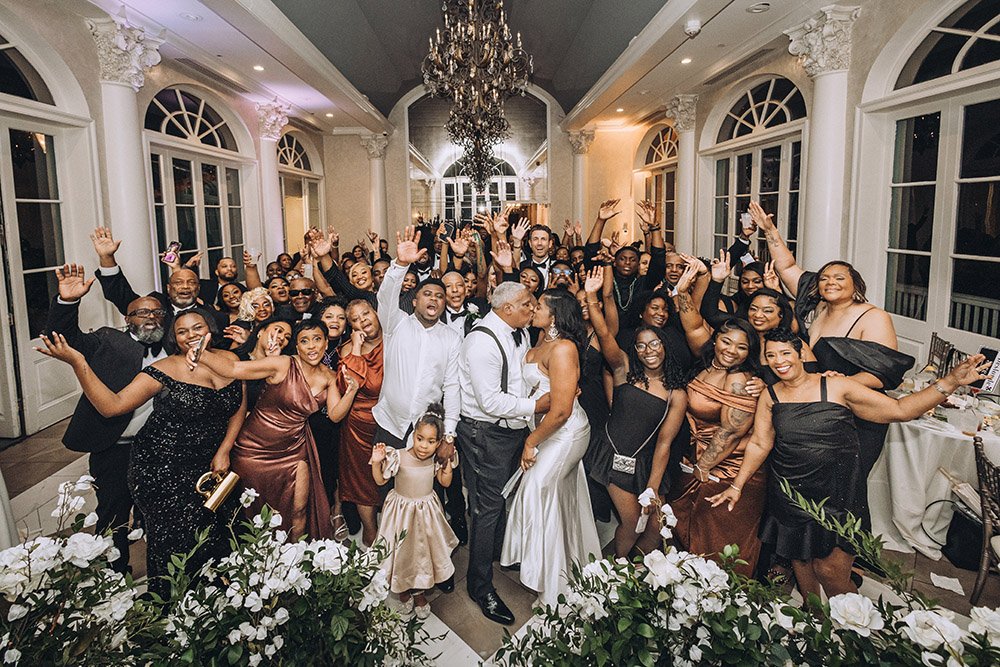The bride and groom kiss at the center of a joyful group portrait as guests raise their hands in celebration inside the ballroom.