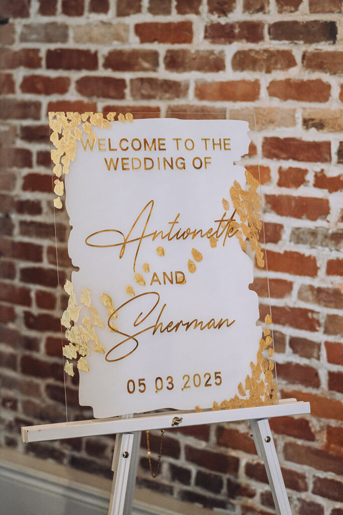 A modern acrylic welcome sign with gold lettering welcomes guests to the wedding of Antionette and Sherman against an exposed brick backdrop.