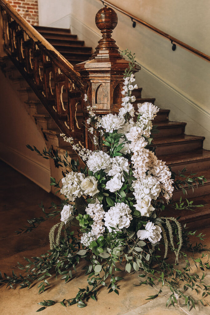 A lush white floral arrangement with greenery displayed beside a historic wooden staircase inside the venue.