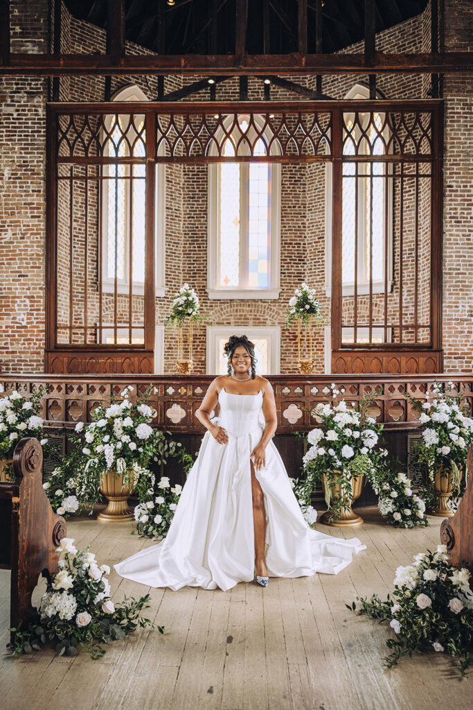 The bride stands centered at the altar in a strapless gown, framed by white florals and exposed brick walls inside the church.