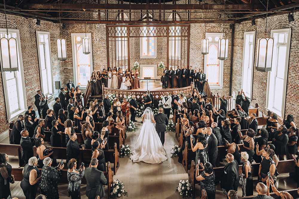 A wide view of the wedding ceremony inside a brick church, with the bride and groom standing at the altar surrounded by seated guests.