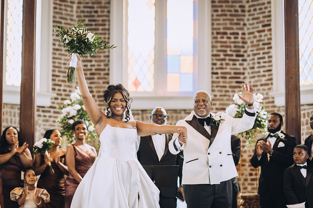 The bride raises her bouquet in celebration while holding hands with the groom at the altar, surrounded by applauding family and guests.