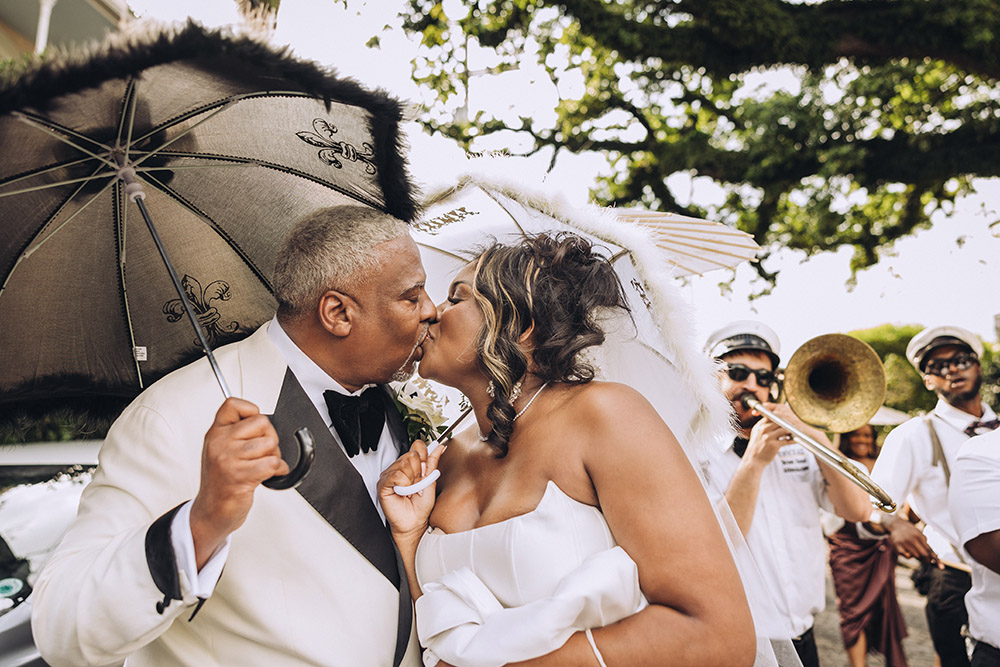 The bride and groom share a kiss beneath lace umbrellas during a lively New Orleans second-line parade with musicians behind them.