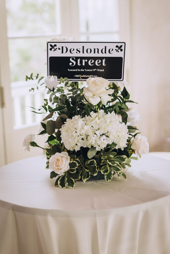 A black and white Deslonde Street table sign sits atop a white floral centerpiece with roses and greenery.