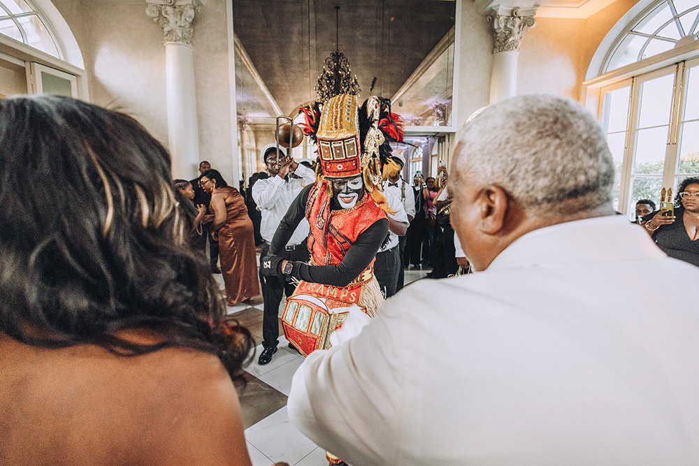 A Mardi Gras Indian performer dances through the crowd during the reception, surrounded by guests inside a grand New Orleans venue.