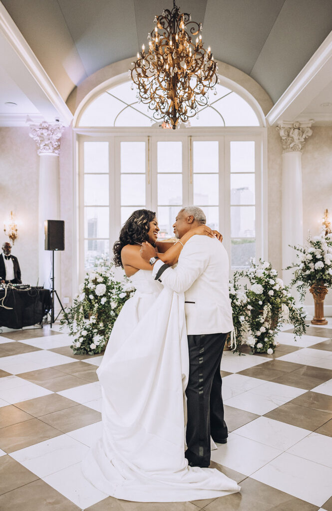 The bride and groom share their first dance beneath an ornate chandelier, surrounded by lush white florals and tall windows.