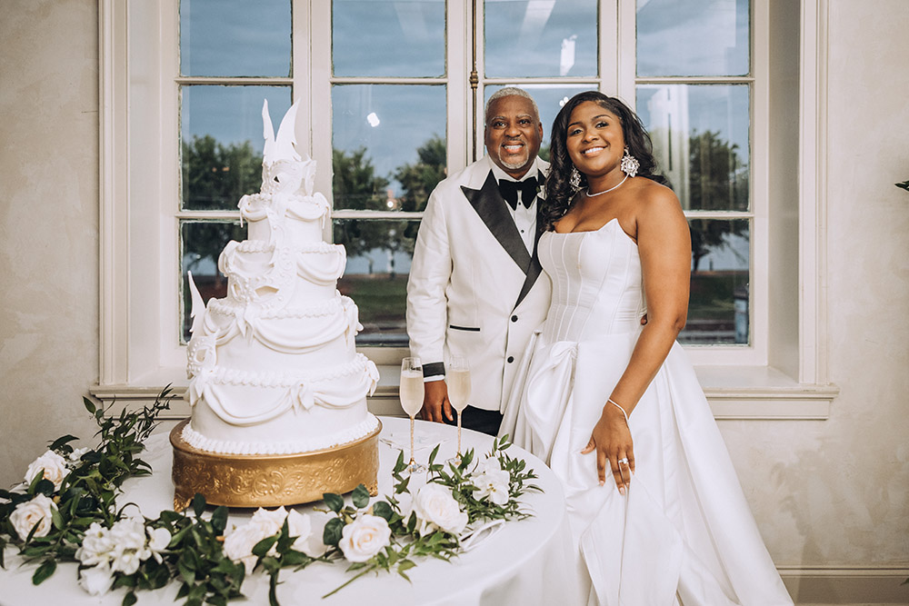 The bride and groom pose beside a sculpted white wedding cake with champagne glasses and greenery accents.