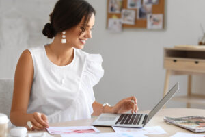 Smiling woman in a white blouse working at a laptop with papers spread across a light-filled workspace.
