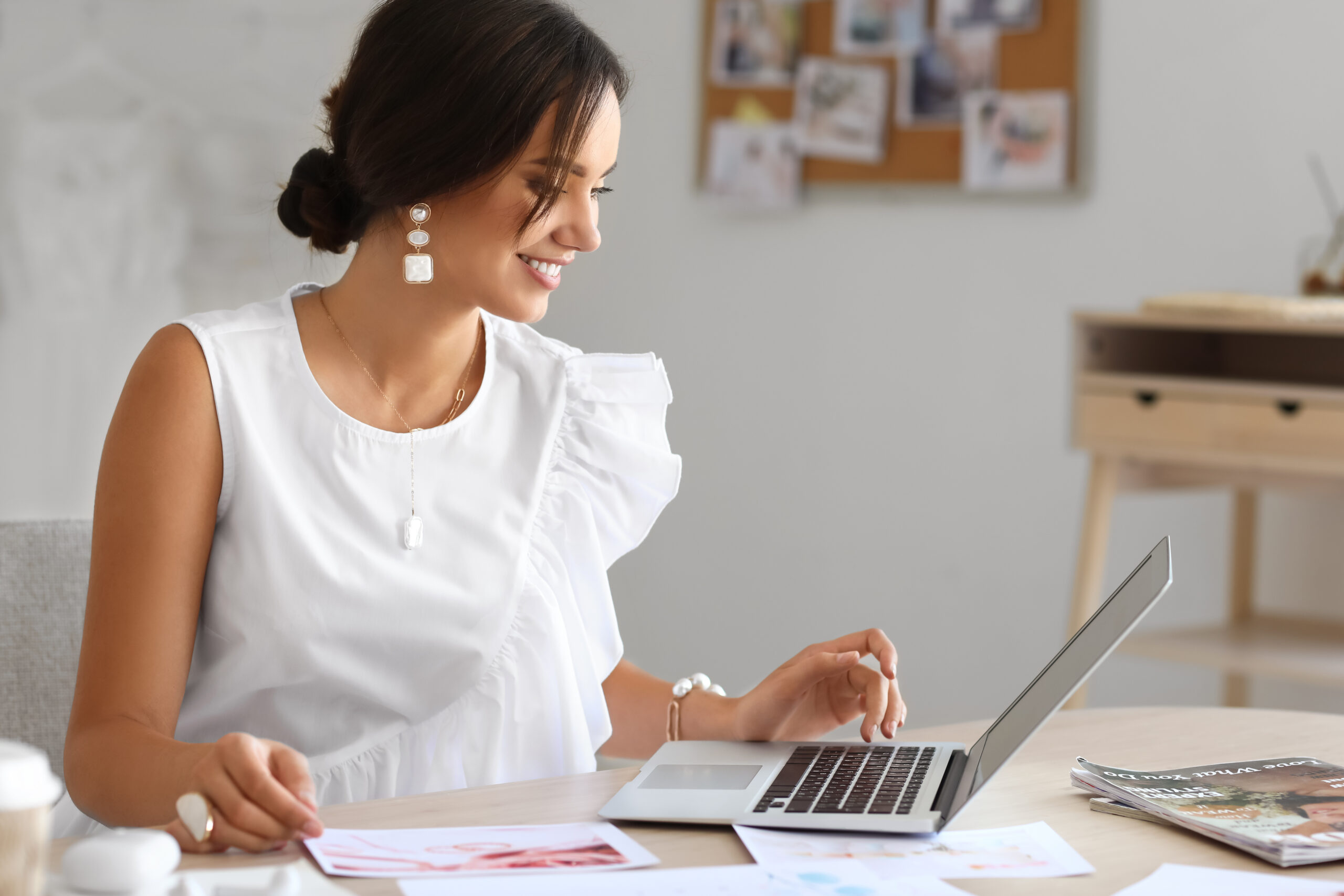 Smiling woman in a white blouse working at a laptop with papers spread across a light-filled workspace.