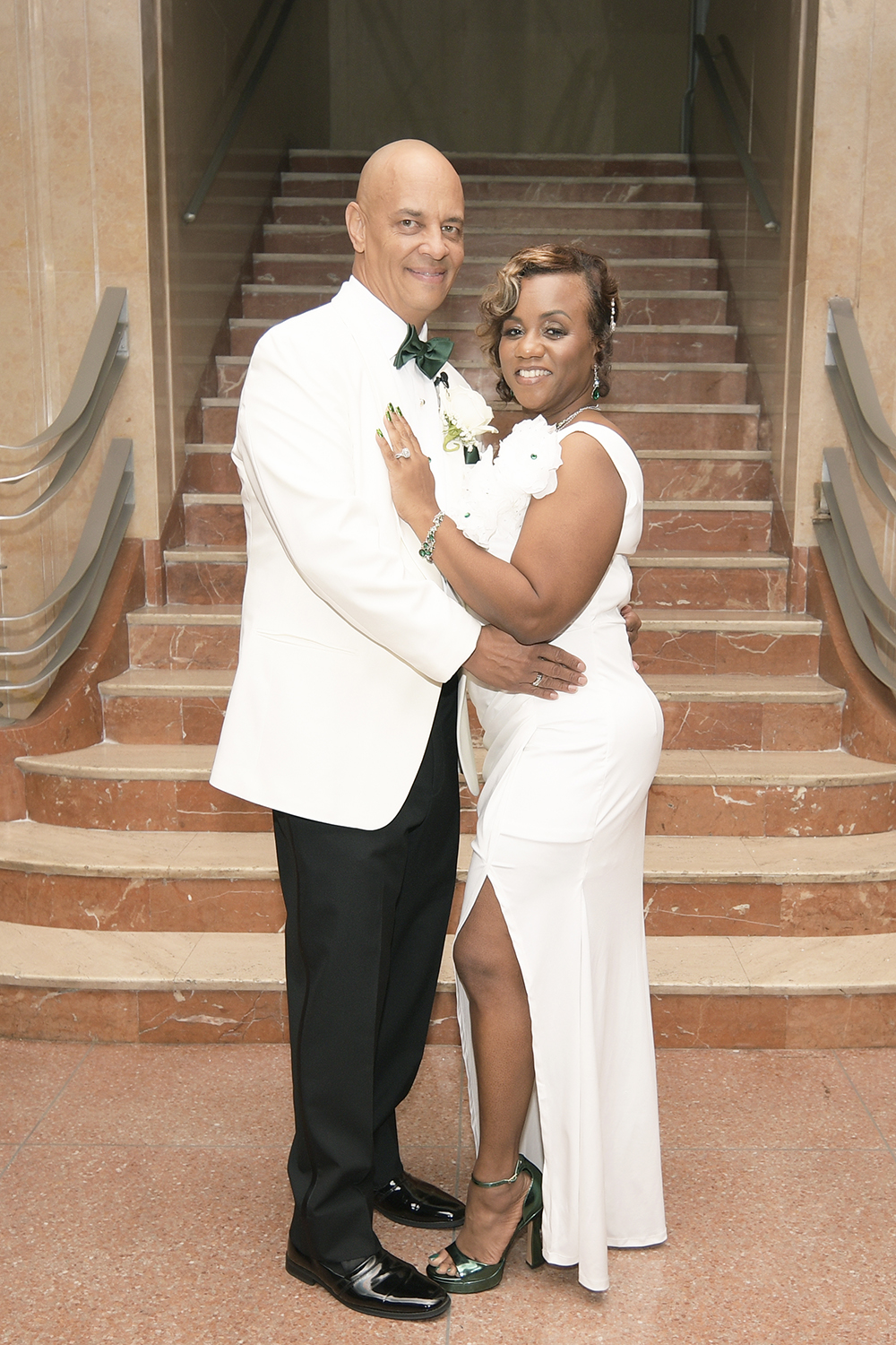 Bride and groom pose together on a marble staircase, dressed in white and black formal wedding attire.