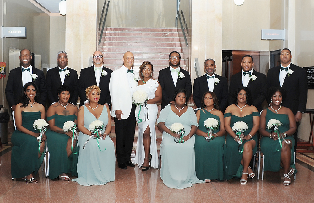 Bride and groom pose with their wedding party on a grand staircase, with bridesmaids in green dresses and groomsmen in black tuxedos.