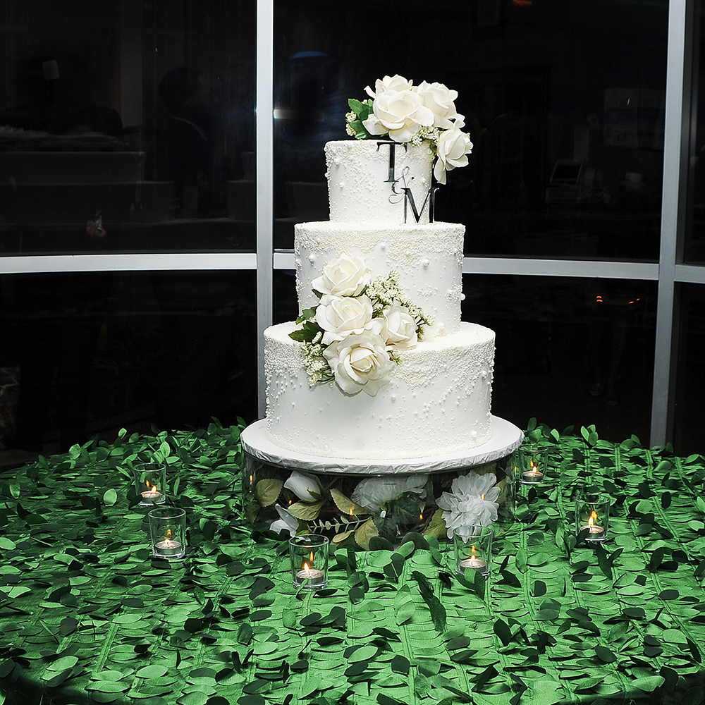 Three-tier white wedding cake decorated with white florals, displayed on a greenery-covered table with candles.