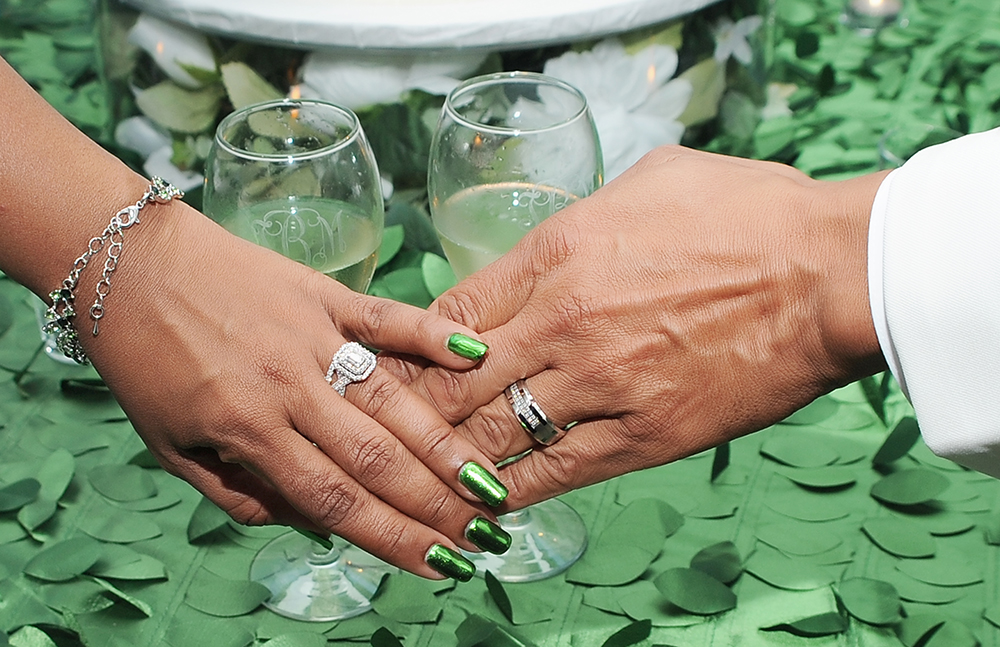 Close-up of the bride and groom’s hands showing wedding rings as they hold champagne glasses on a greenery-covered table.