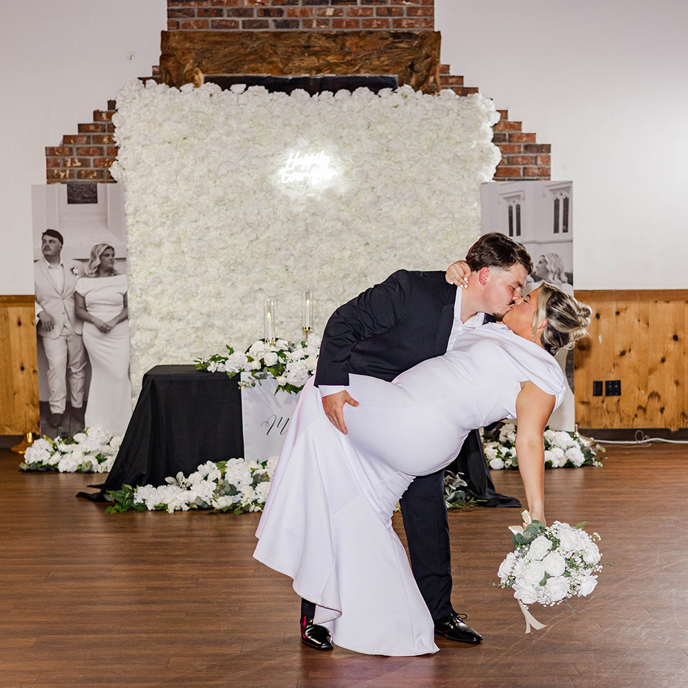 Bride and groom share a dipped kiss on the dance floor in front of a white floral backdrop.