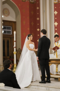The bride smiles at the groom during the ceremony as they stand together at the altar inside a red and gold cathedral interior.