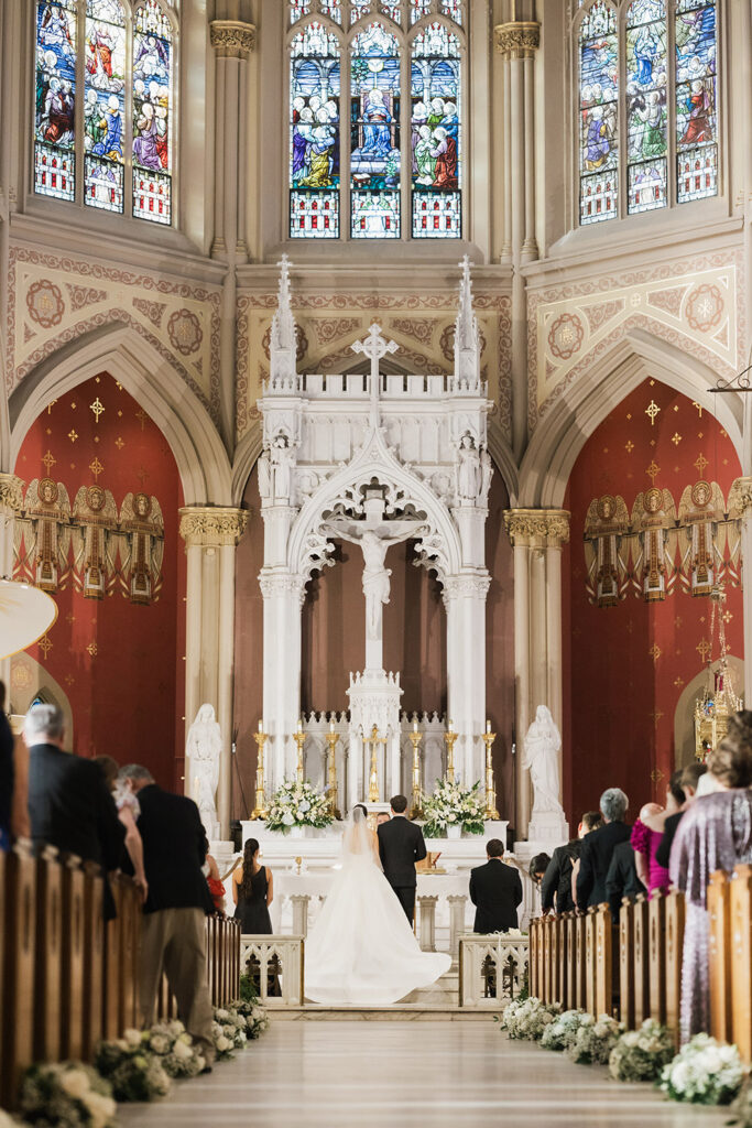 A wide view of the wedding ceremony at the altar, with the bride and groom framed by stained glass windows and ornate church architecture.