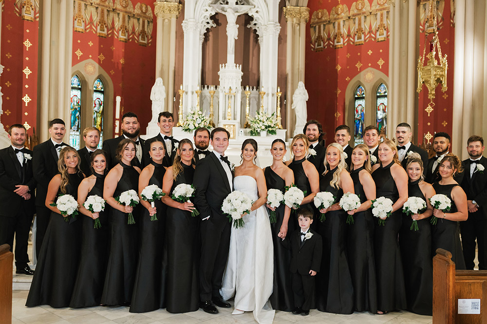 A formal wedding party portrait at the altar, with the bride and groom surrounded by bridesmaids in black gowns and groomsmen in tuxedos.