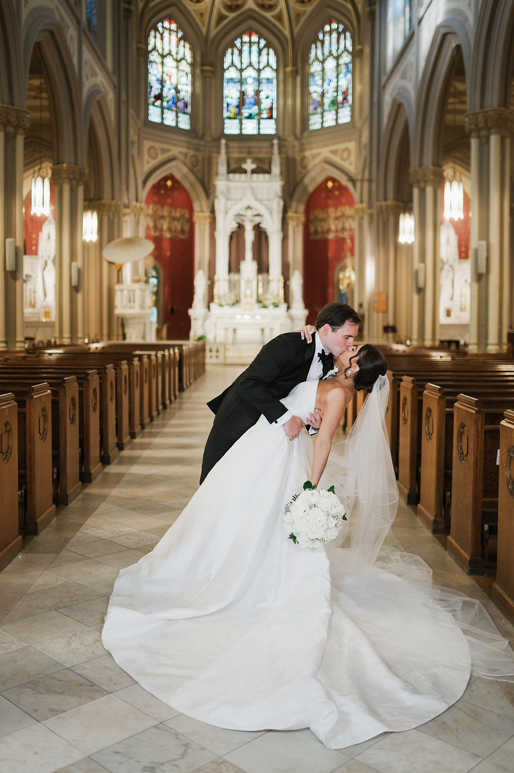The bride and groom share a dramatic dip and kiss in the center aisle of a cathedral, framed by stained glass windows and wooden pews.