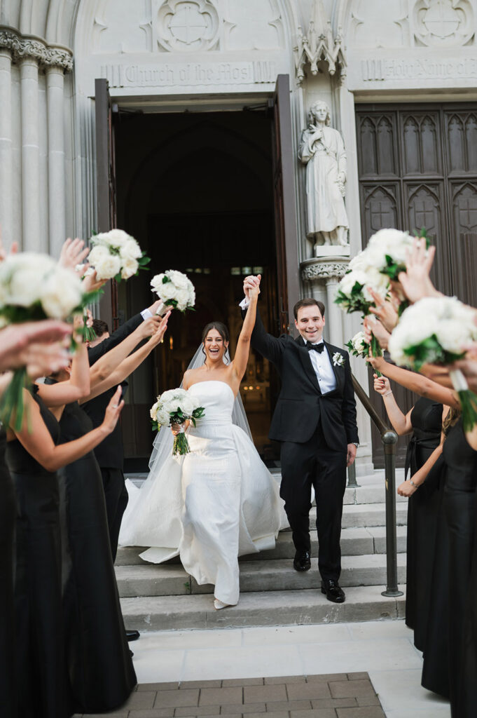 The newlyweds exit the church hand in hand as the wedding party cheers and raises white floral bouquets outside the historic entrance.
