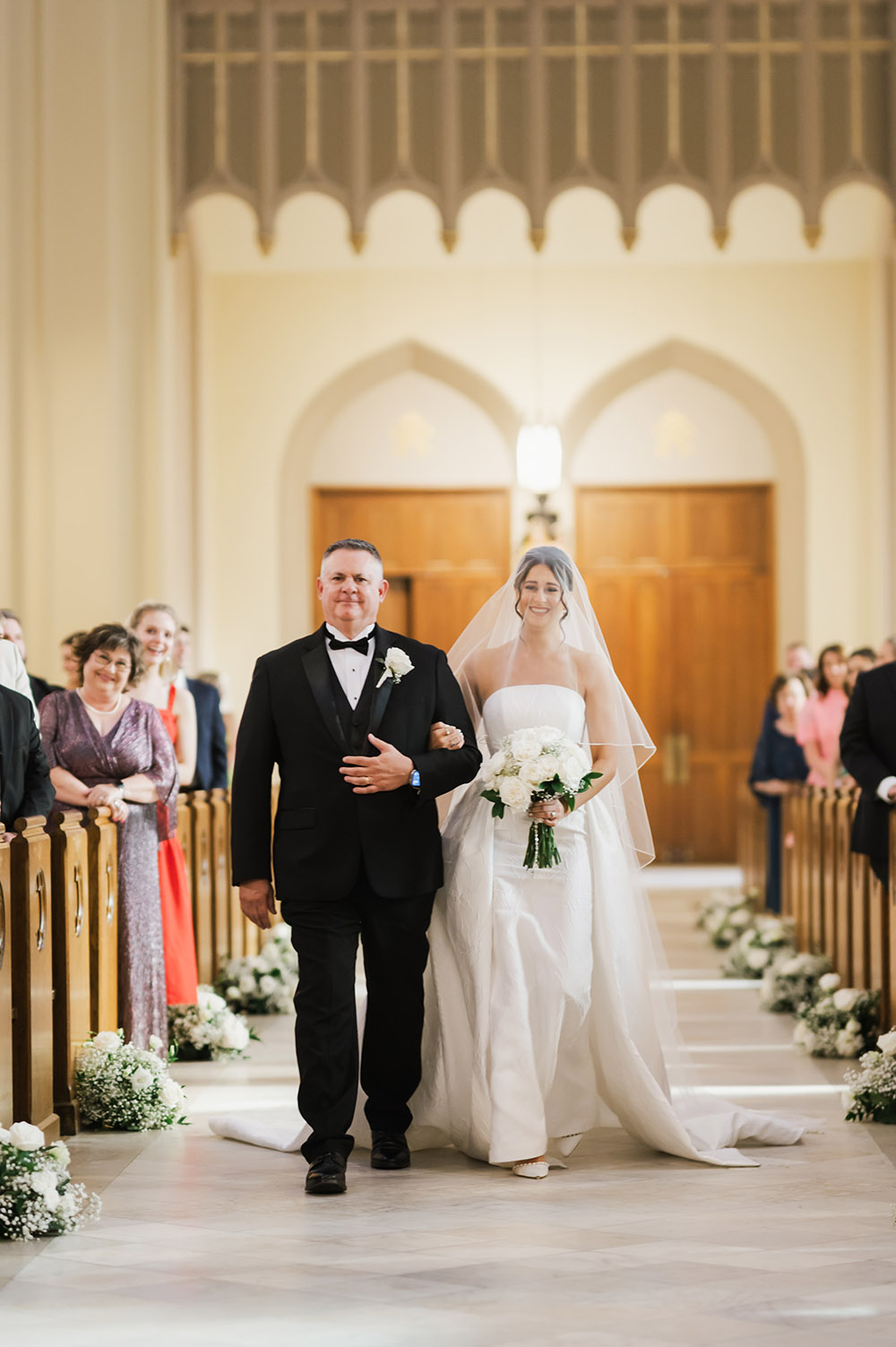 The bride walks down the aisle with her father inside a grand cathedral, holding a white bouquet as guests look on from wooden pews.