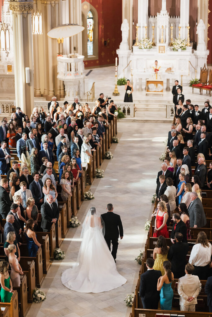 An overhead view of the bride and her father processing down the center aisle of a cathedral church, surrounded by seated guests and floral aisle arrangements.