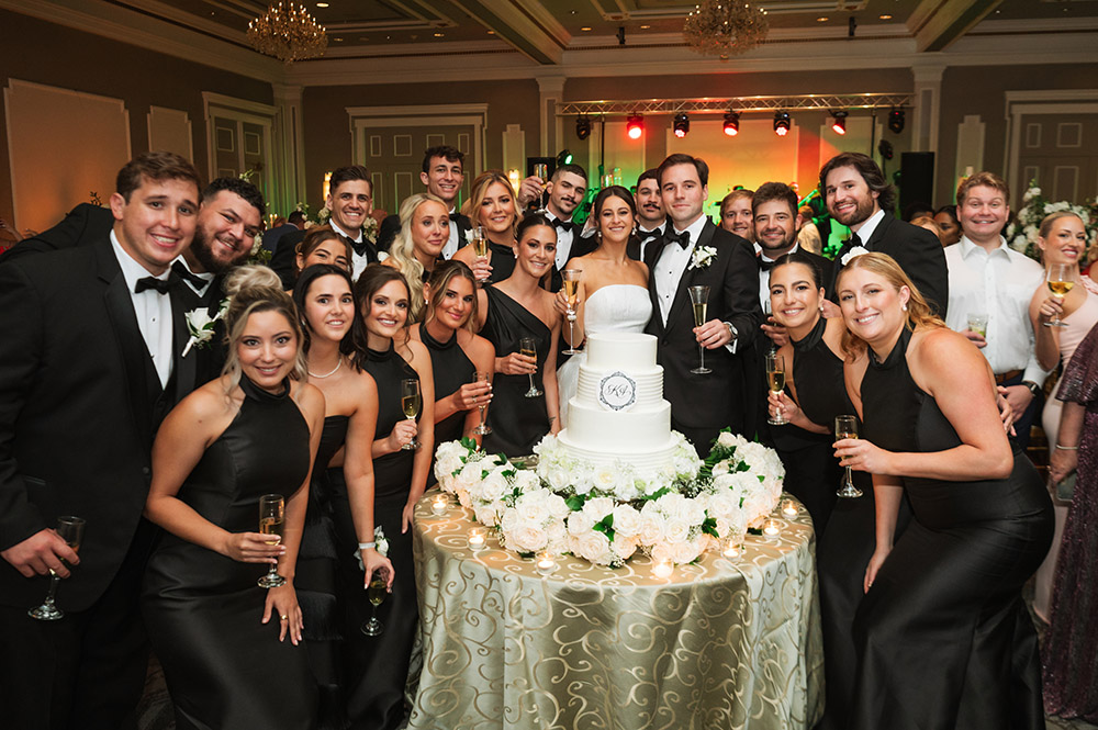 The bride and groom pose with their wedding party around a tiered white cake, raising champagne glasses in celebration.
