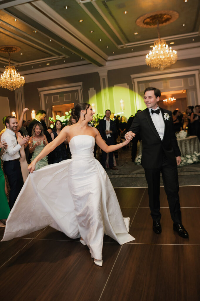 The bride twirls onto the dance floor holding hands with the groom as guests cheer during the wedding reception.