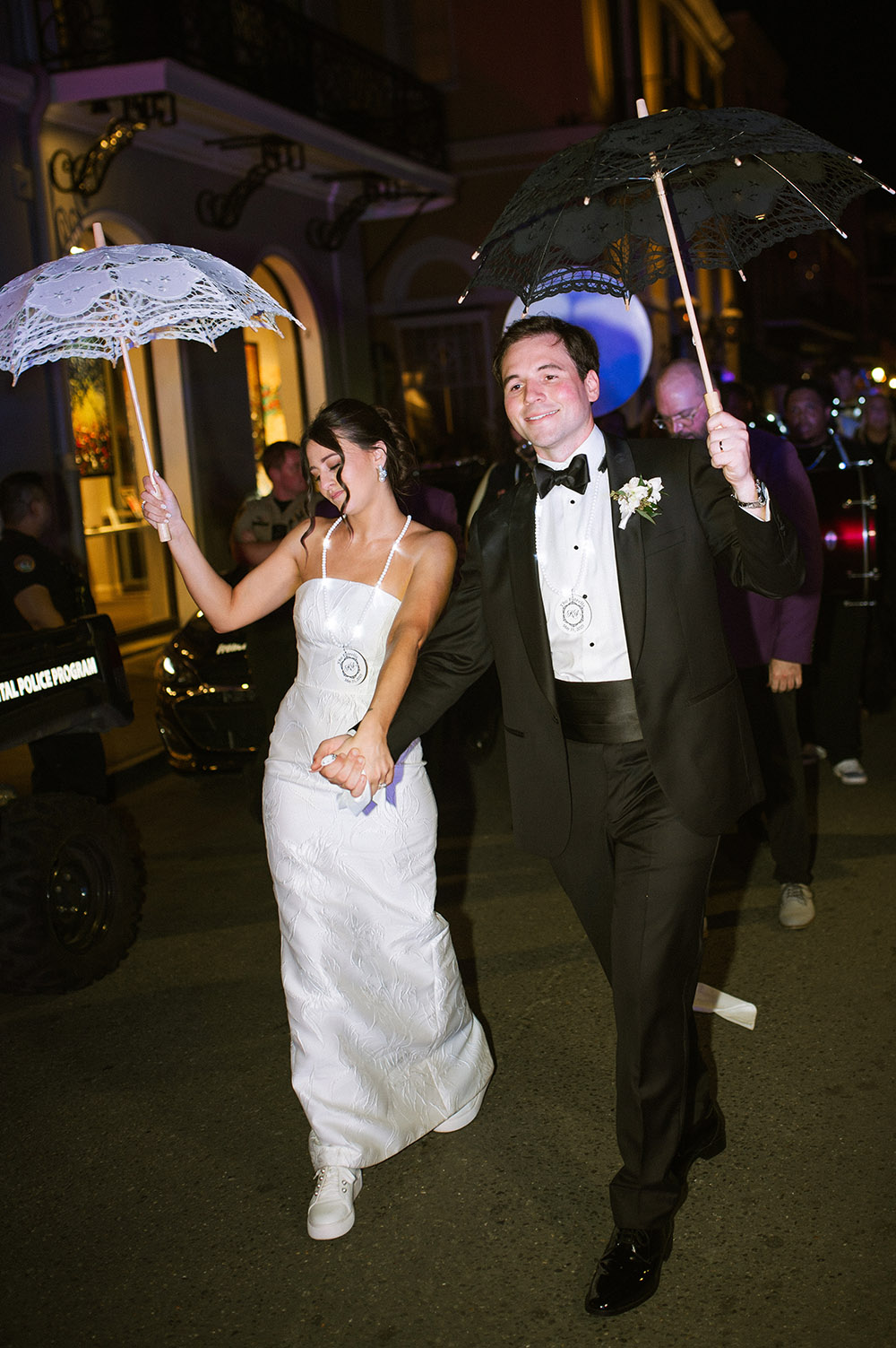 The bride and groom lead a nighttime second-line parade, holding lace umbrellas and wearing beads as guests follow behind.