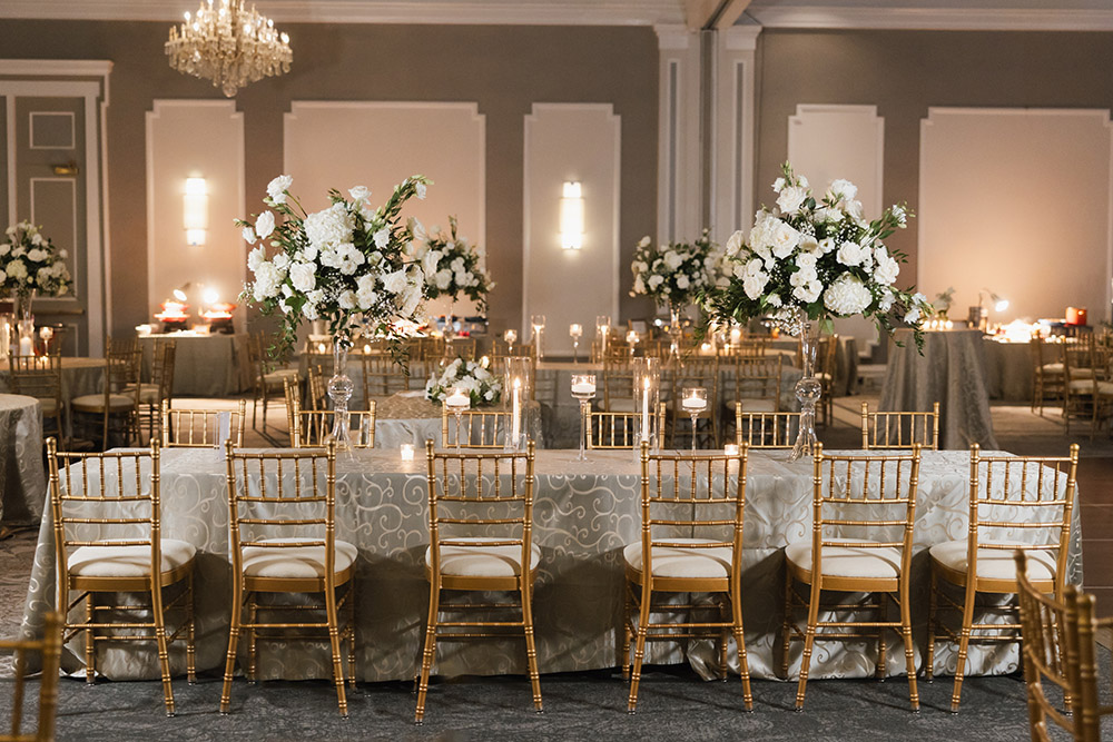An elegant reception table set with gold chiavari chairs, soft candlelight, and tall white floral centerpieces beneath crystal chandeliers.