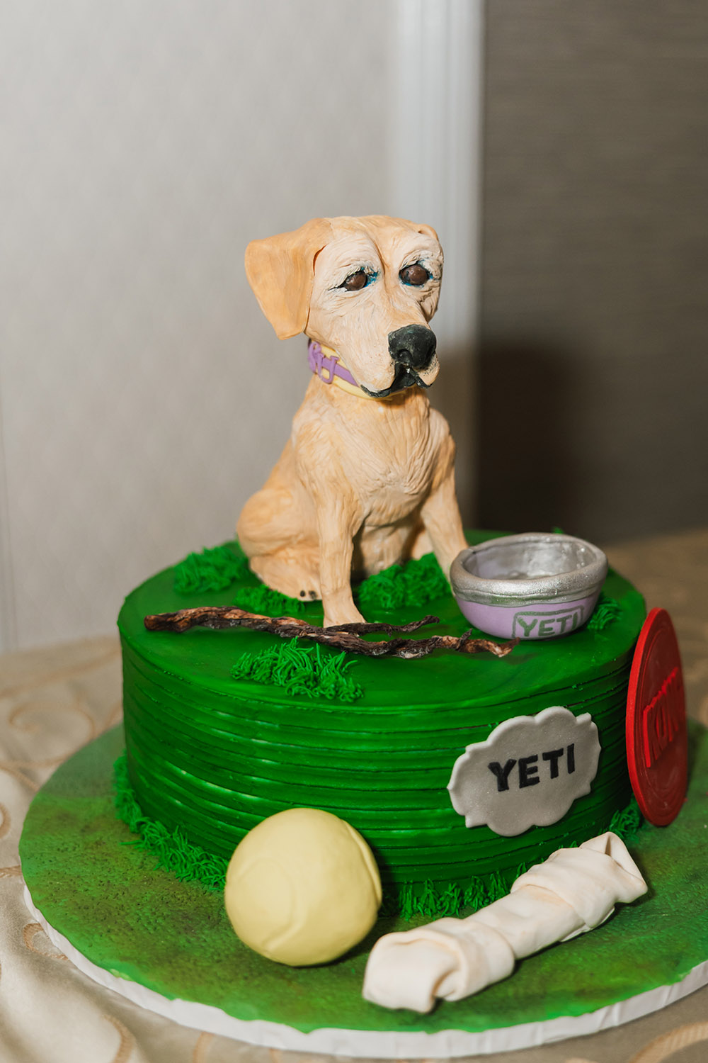 A playful groom’s cake featuring a sculpted dog topper with a YETI bowl, tennis ball, and bone on a green frosted base.
