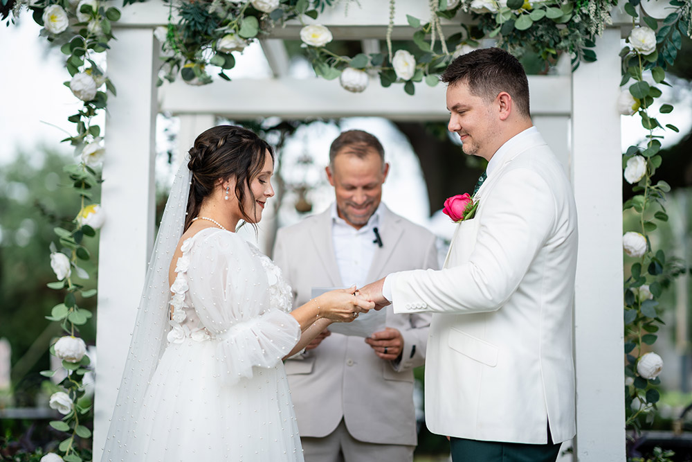 The bride and groom exchange rings during an outdoor ceremony beneath a floral-covered arbor as the officiant looks on.