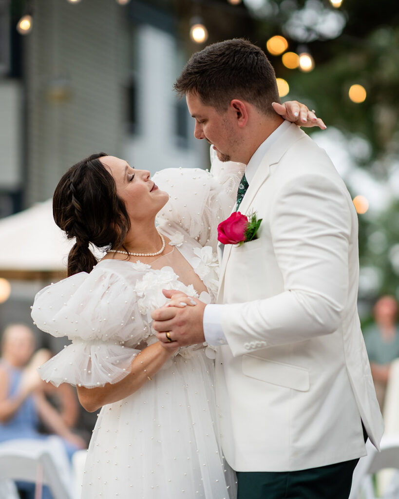 The bride and groom share their first dance outdoors beneath string lights, holding hands and smiling softly at one another.