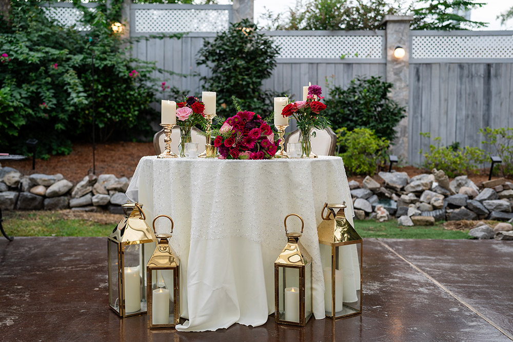 Outdoor sweetheart table styled with gold lanterns, ivory linens, candles, and deep pink floral arrangements set against a garden backdrop.