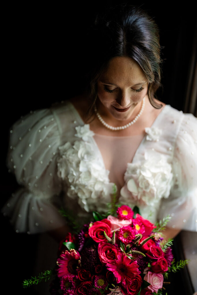 Bride portrait holding a vibrant pink and burgundy bouquet, softly lit with a dark background highlighting her textured wedding gown.
