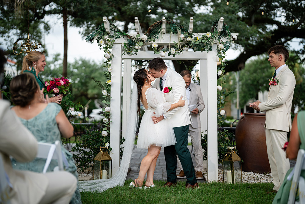 Outdoor ceremony kiss beneath a white pergola adorned with greenery and white flowers, surrounded by guests as the couple shares their first kiss.