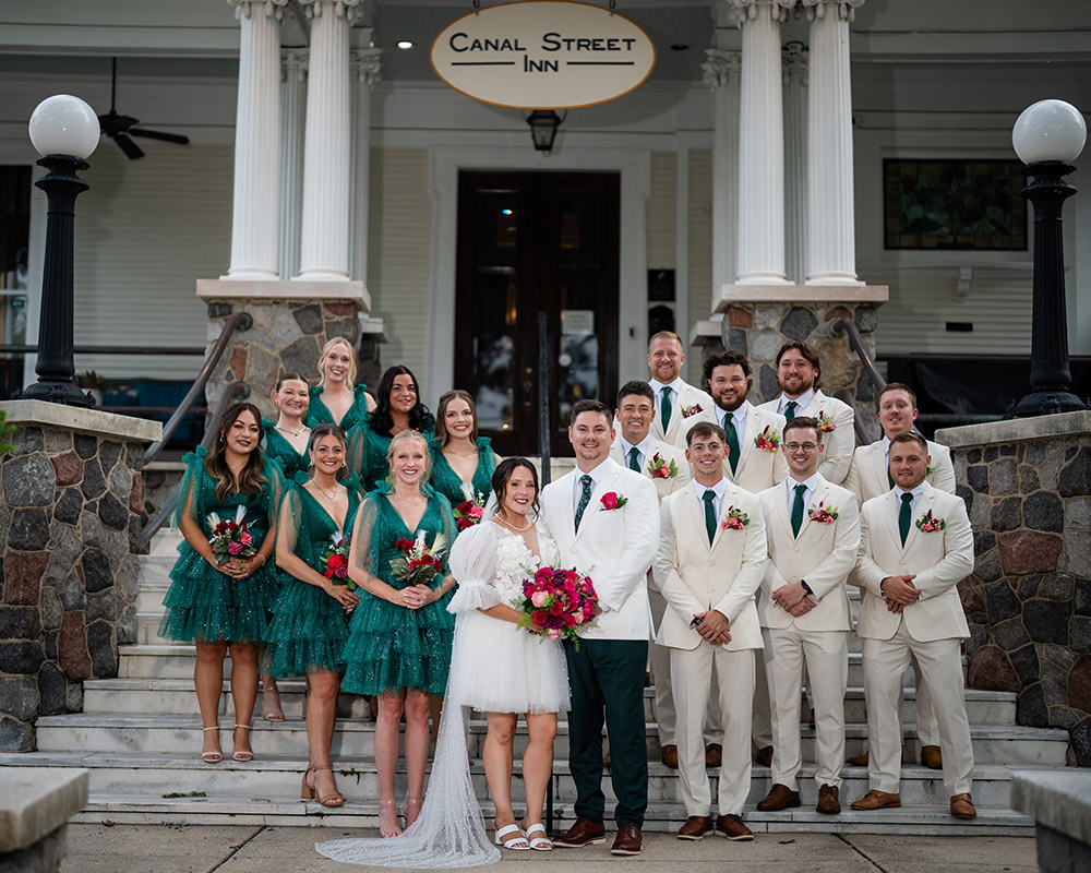 Wedding party group portrait on the steps of Canal Street Inn, featuring the bride and groom with bridesmaids in green dresses and groomsmen in light suits.