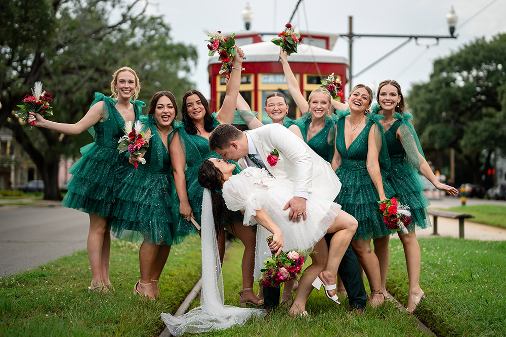Bride is dipped by the groom while bridesmaids in emerald green dresses cheer and raise bouquets along a New Orleans streetcar line.