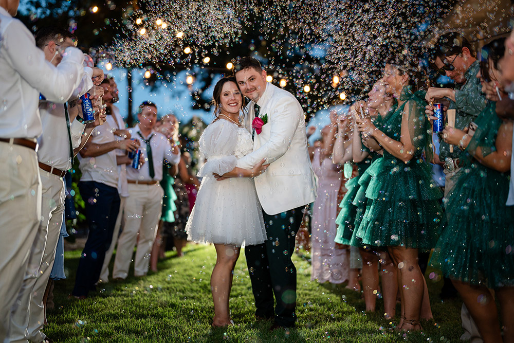 Bride and groom stand arm in arm beneath floating bubbles, smiling during their festive outdoor wedding send-off.