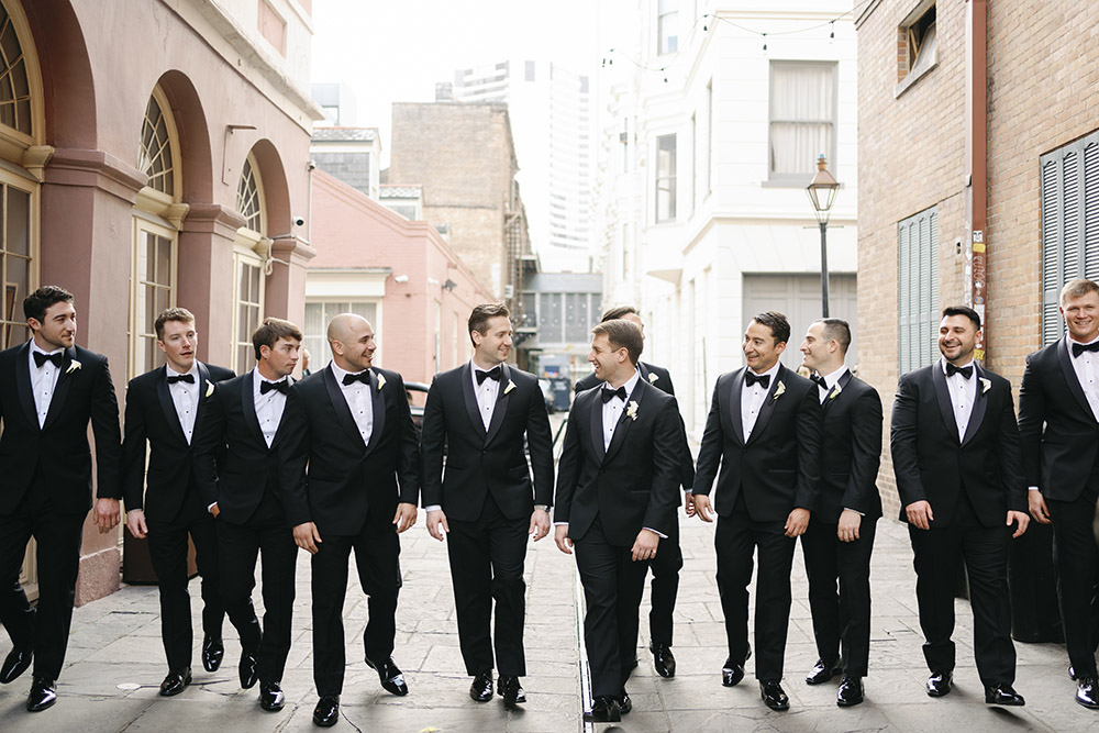 Groom and groomsmen walking together in black tuxedos through a historic city street on the wedding day.